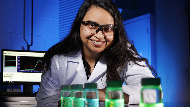 Shudipto Dishari in a lab coat with glowing green bottles, computer screen displaying graphs in the background.