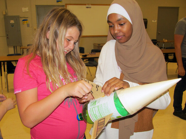 Two individuals collaborate on building a model rocket in a classroom setting.