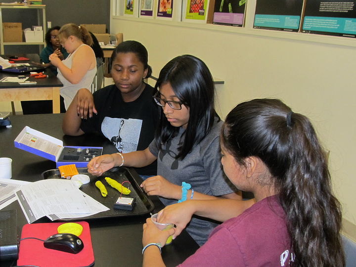 Students collaborating at a classroom table with tools, papers, and educational posters on the wall.