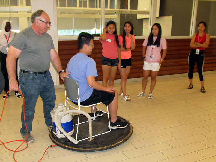 Man demonstrates a hovercraft chair to a seated individual as a group of people looks on indoors.